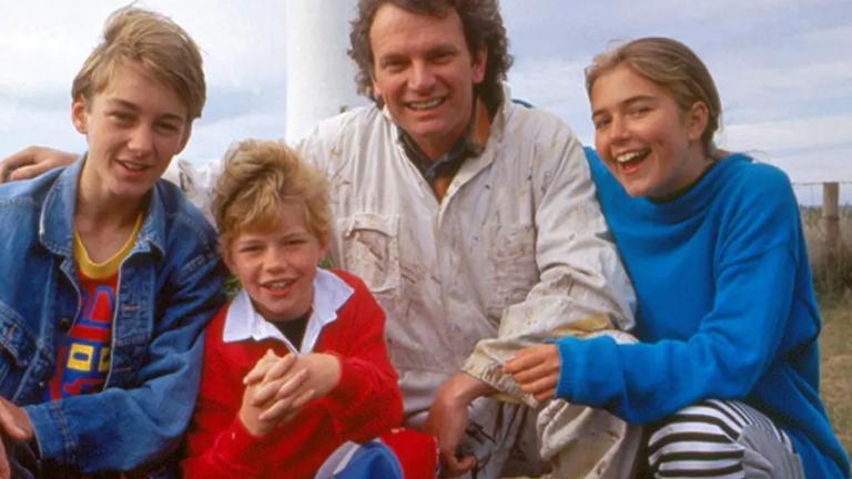 A family of four - two boys, a girl and their dad, kneeling with their arms around each other and smiling at the camera. You can see part of a lighthouse in the background. 