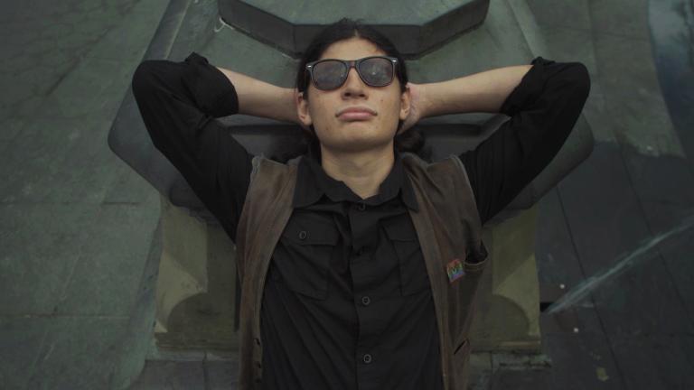 A young man pictured from the waist up wearing dark sunglasses, leaning back with his arms raised and hands folded behind his head.