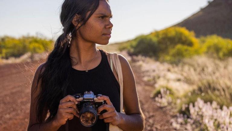 A teenage First Nations girl standing on an outback dirt road holding a camera and looking into the distance.
