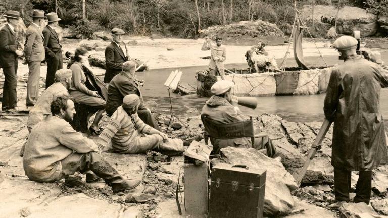 A film crew and actors on a rocky beach filming a scene in which two men dressed as convicts and a woman are on a makeshift boat.