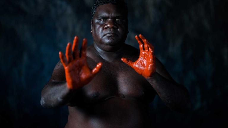 A First Nations man pictured from the waist up with his hands out in front of him. His hands are covered in red pain and he is looking directly at the camera. He is pictured against a dark background. 