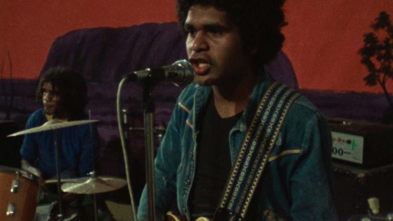 A First Nations man playing guitar on stage with a rock band.