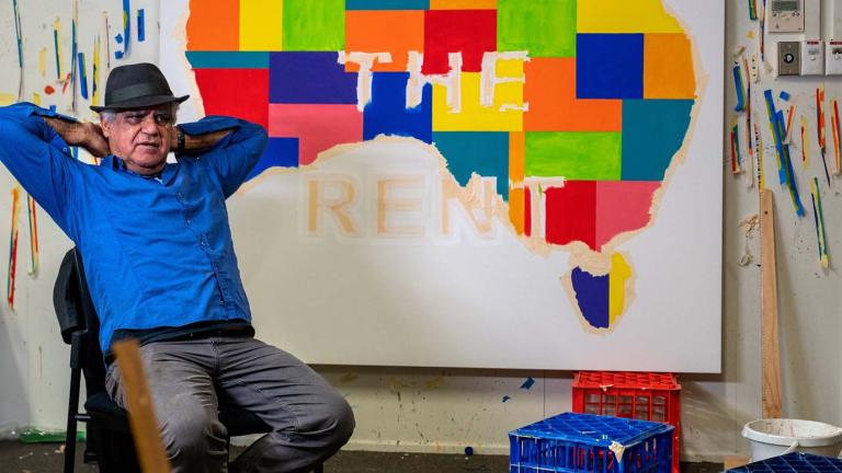 Artist Richard Bell seated in his studio in front of one of his works: a coloured Indigenous map of Australia with the words PAYE THE RENT across it
