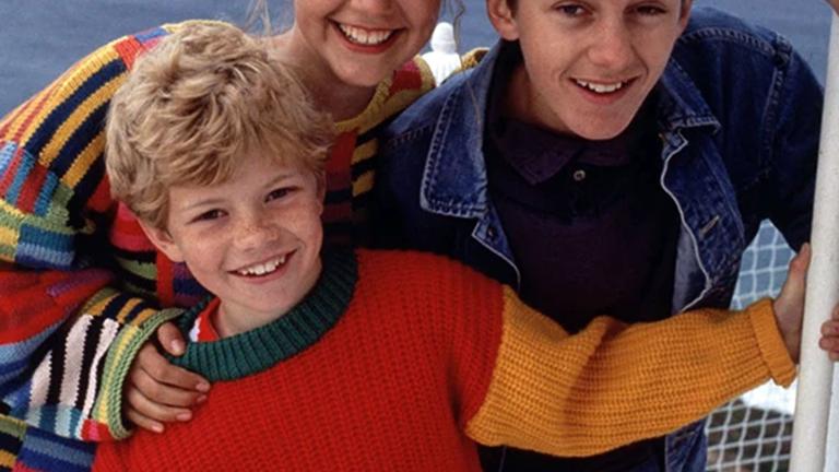 Three white children, two boys and a girl, laughing and smiling together on a boat at sea. The youngest boy and girl are wearing colourful jumpers while the older boy wears a denim jacket.