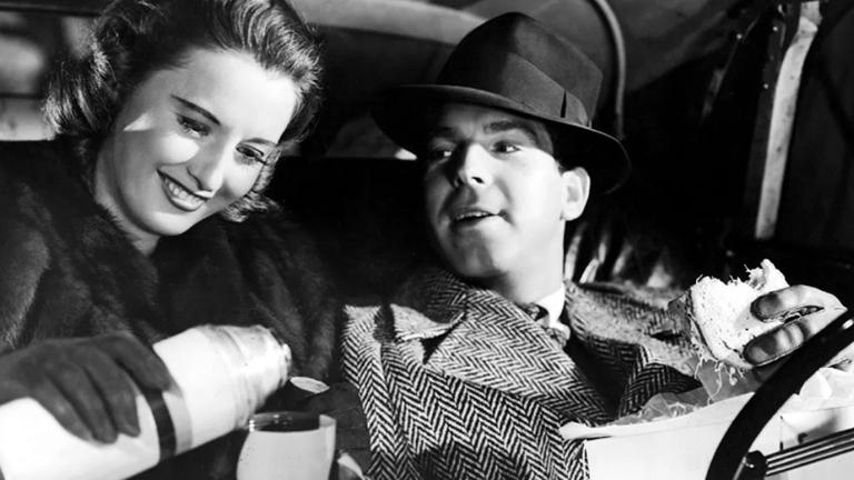 A woman and man sitting side by side. They are both wearing warm coats and gloves. The woman is pouring a drink from a thermos into a cup and smiling and the man is looking at her while holding a sandwich in one hand.