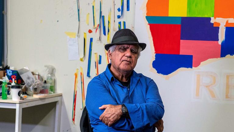 Artist Richard Bell seated in his studio in front of one of his works: a coloured Indigenous map of Australia with the words PAYE THE RENT across it