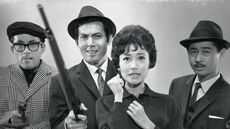 Four Japanese people - 3 men and a woman - pictured standing side by side looking towards the camera. The men are all wearing suits and hats and holding firearms.