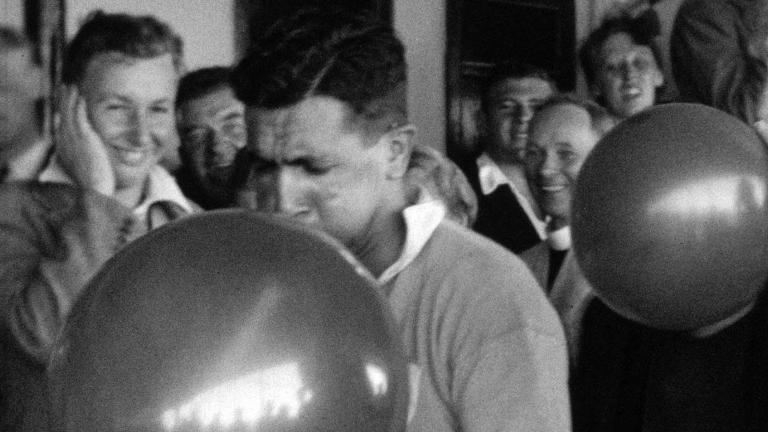 Two men blowing up balloons while a group of other men standing in the background are watching