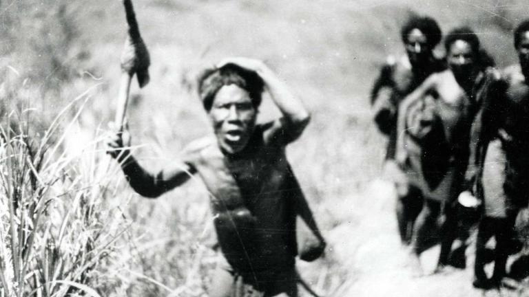 A group of four Indigenous men walking through a grassy area. One of them is holding a tool of some description in one hand, with his other hand on his head.