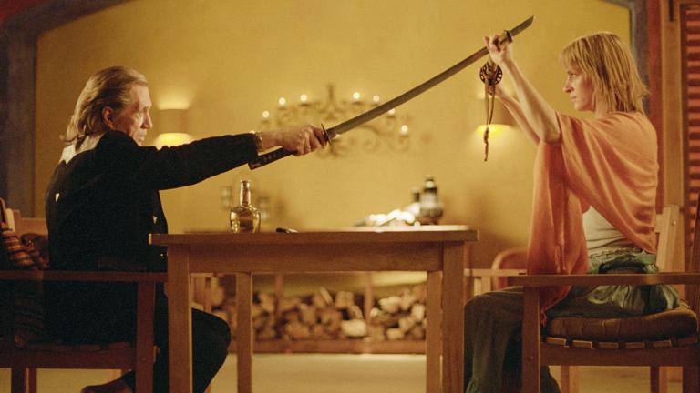 A man and woman seated at each end of a dining table. The man is holding a samurai sword above the woman's head and she is blocking it with another sword, in a scene from Kill Bill: Volume 2.