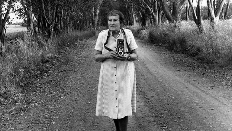 Wide shot of an older woman standing in the middle of a dirt road that is lined by trees. She is holding an old camera.