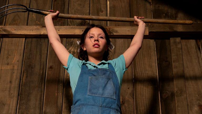 An angry-looking teenage girl standing in what looks to be a barn, is holding a pitchfork above her head.