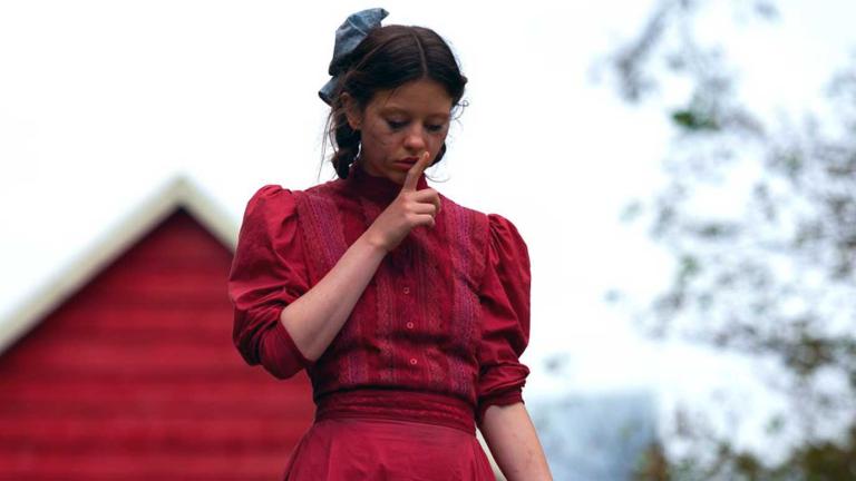 A teenage girl pictured from the waist up, standing with her head bowed and an index finger against her lips in a 'shushing' gesture. There are trees and the roof of a house behind her.