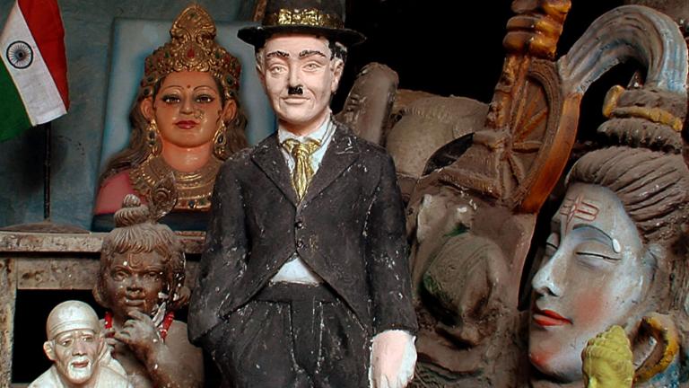 A statuette of silent film star Charlie Chaplin sitting on a shelf net to some Indian statues and trinkets and an Indian flag.