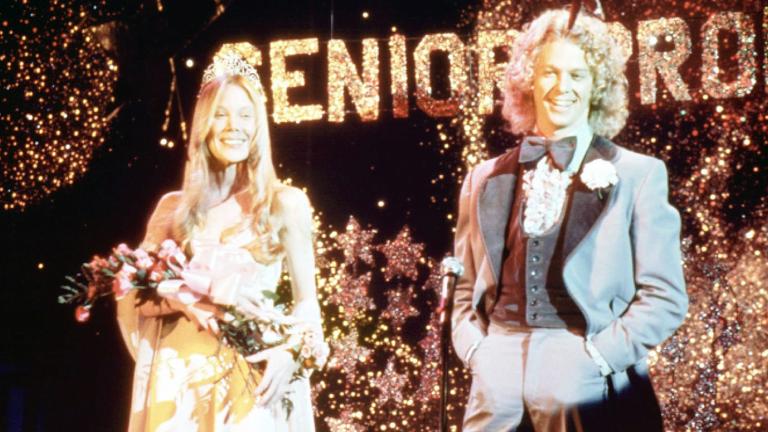 A teenage girl and boy at Senior Prom. The girl is wearing a tiara and holding a bunch of flowers. The boy is wearing a tuxedo. They are both smiling.