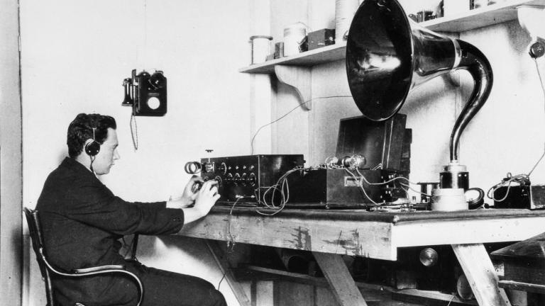 A man sitting on a desk with headphones on. There is radio and other audio equipment on the desk. c1920s