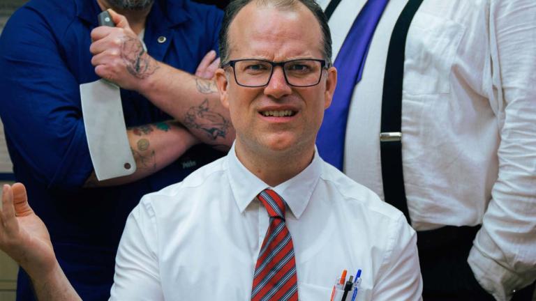 A seated white man in a white business shirt and tie and wearing glasses looks exasperated and holds up one hand. Two men are standing close behind him.