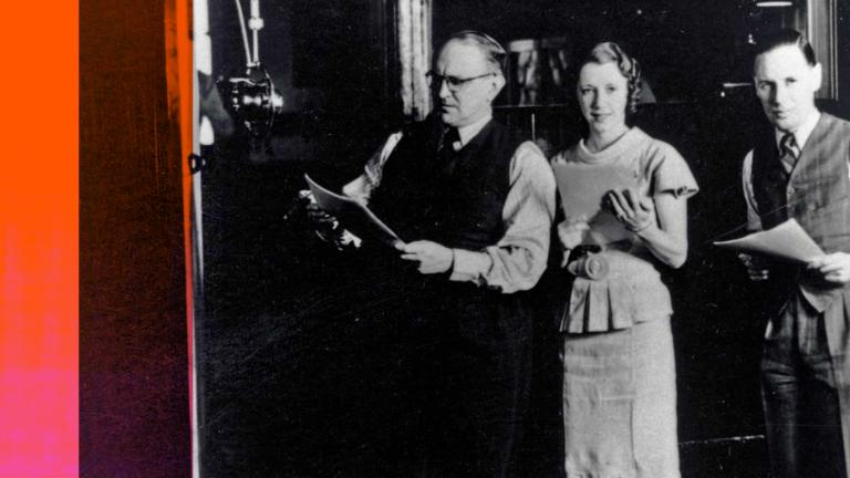 A man and two women in a recording studio standing next to a big radio microphone. They are holding scripts in their hands.