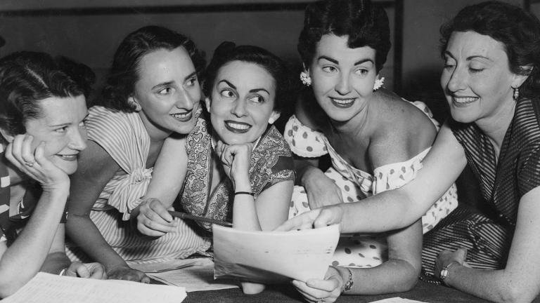 Five women leaning against a desk looking at a sheet of paper, they are all smiling. 