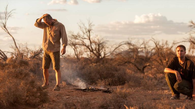 Guy Pearce and Robert Pattinson by a smouldering bushfire in an outback setting either at dusk or early dawn. Pattinson is crouching while Pearce stands apart, looking down, with hand on his head