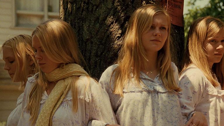Four young women with long blonde hair wearing pale nighties and standing outside around a tree during the daytime, with mournful expressions