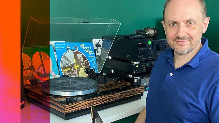 NFSA curator Simon Smith, seated and looking up at the camera. He is sitting at a desk with a turntable and vinyl records on it.