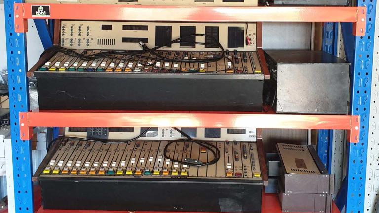 A shelving unit holding a number of radio mixing consoles.