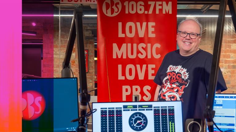 A man stands behind a radio station mixing desk and monitor smiling. He stands in front of a banner that says "106.7FM Love Music Love PBS".