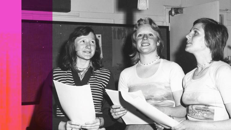Three women holding scripts in a radio studio are standing around a microphone suspended above them