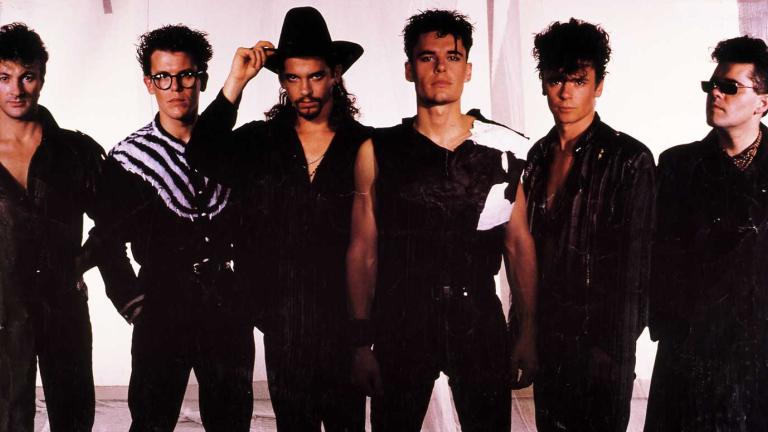 Six members of the band INXS, pictured standing side by side, dressed in black and posing for a publicity image. 