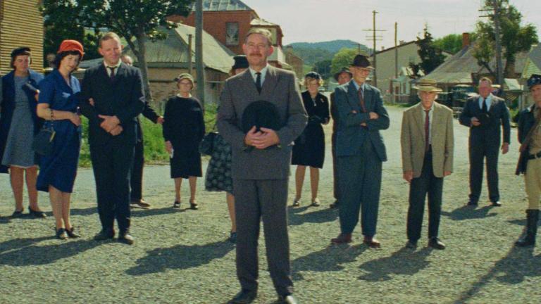 Wide view of a street in a country town. Many of the townspeople are standing in the street looking in the direction of the camera. 