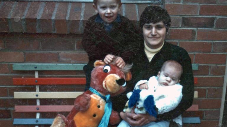 A woman sitting on a bench holding a baby with a young boy behind her.