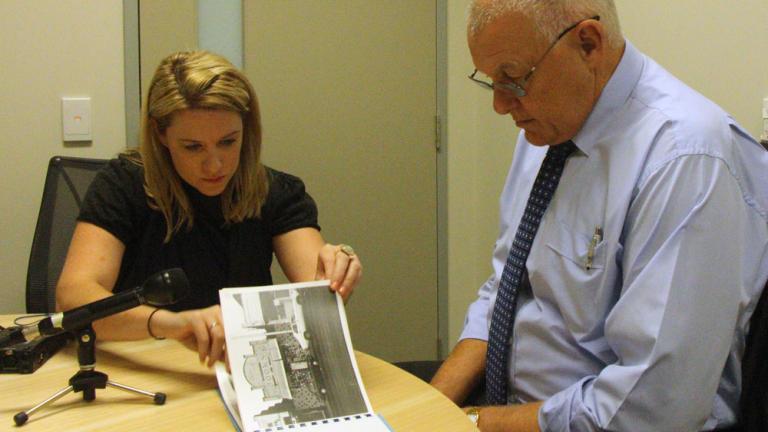 Journalist Rachael Brown at a table interviewing policeman Ron Iddles. There is a microphone and a photograph on the table. 