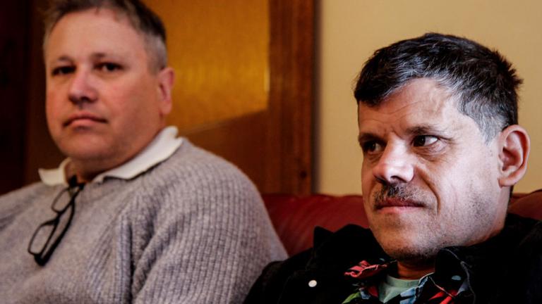 Head and shoulders shot of two men. They are seated, one is looking at the camera and the other straight ahead.