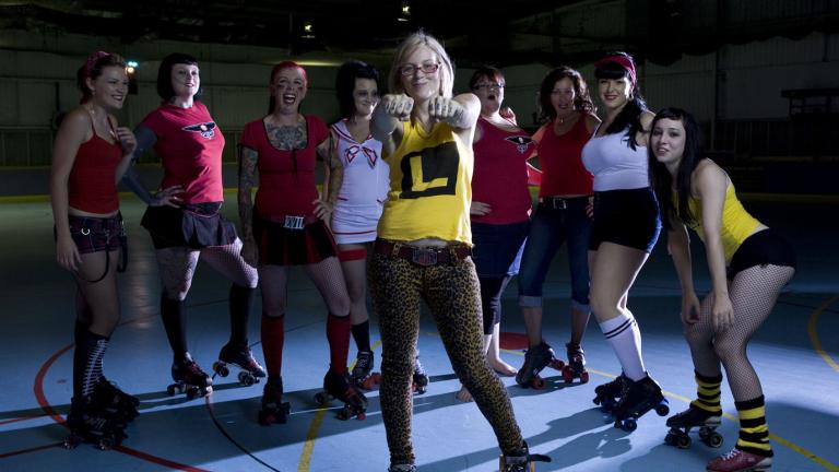 Nine female roller derby players wearing roller skates and standing in a semi-circle, posing. The middle player stands in front of the others and has her arms outstretched and hands bunched into fists.