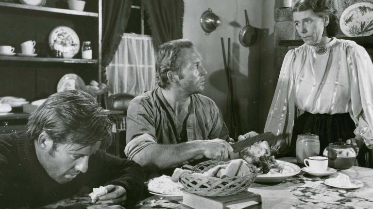 Two tired looking men seated at a table talking to a woman who is standing looking at them