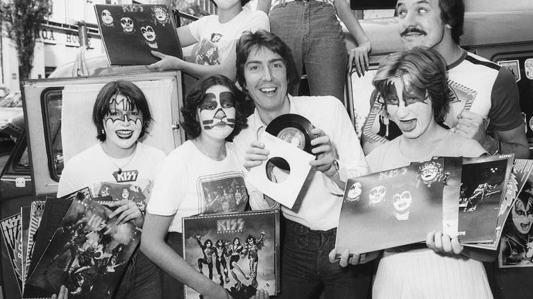 Radio announcer Ron E Sparks surrounded by fans of the band Kiss who have painted faces and are holding up Kiss albums and photos.