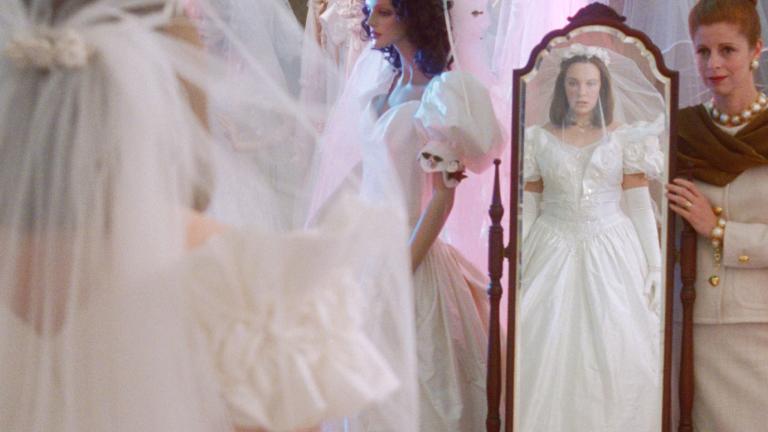 A young woman in a wedding dress looking at herself in a full length mirror while an older woman looks on, in a scene from the film Muriel's Wedding. 