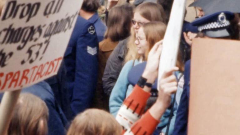 Two women standing in a crowd of protesters and police. A placard near them reads "Drop all charges against the 53".