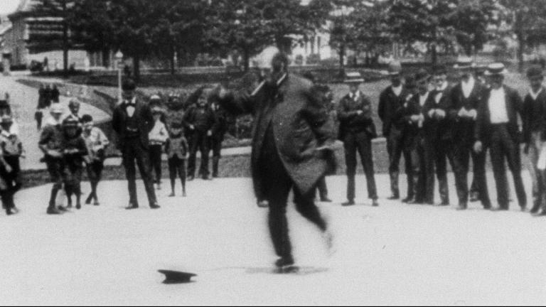 A man skating in a park in front of a crowd of people, circa 1896.