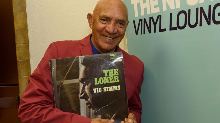 Vic Simms holding up a copy of his vinyl record "The Loner" and standing in front a banner advertising the NFSA's Vinyl Lounge