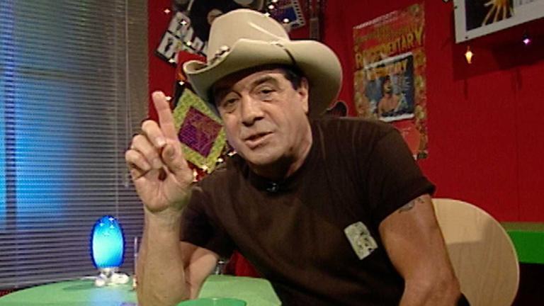 Music television presenter Ian 'Molly' Meldrum seated, wearing a hat, and holding one hand up and pointing his index finger. 