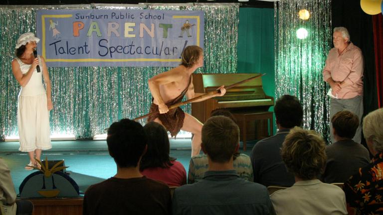 On stage at a school parents' talent show, a woman in a frilly hat and old-fashioned nightgown holds the microphone while a man in a loincloth holds a spear and lunges towards the compere standing in the wings.