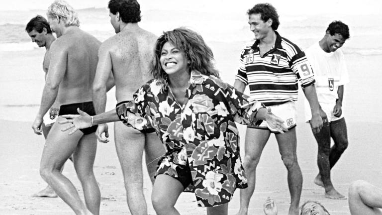 Tina Turner laughing on a beach, surrounded by male rugby league players in swimming costumes.