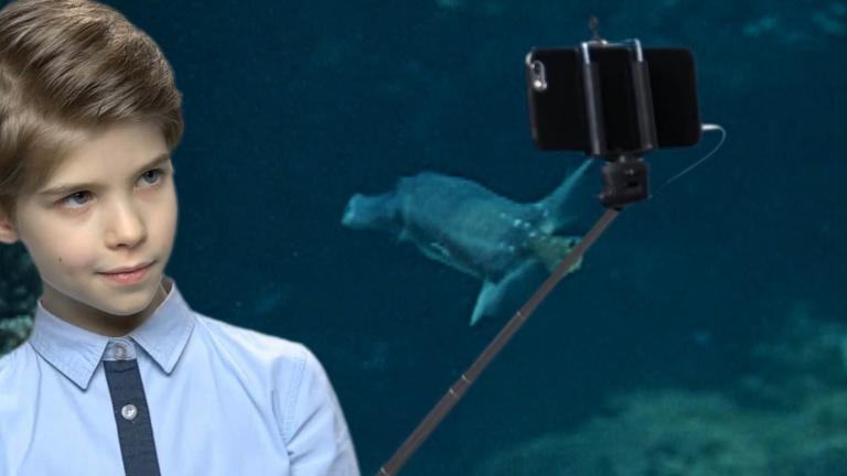 A boy holds a phone on a selfie stick against the backdrop of a video screen showing an underwater scene.