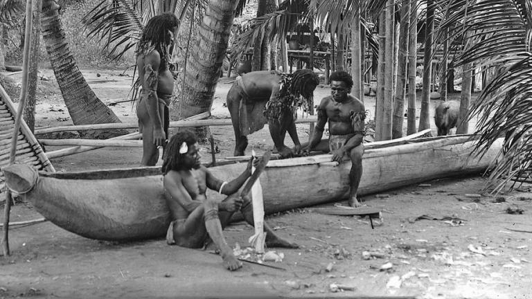 A group of Torres Strait Islander men building a canoe by the water, surrounded by palm trees in the early 1920s.