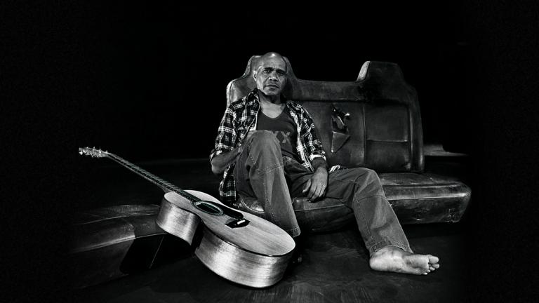 Archie Roach rests against an old car seat in a studio with his guitar resting nearby.