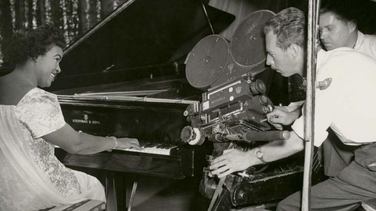 Winifred Atwell playing the piano and smiling while two men film her with a 35mm film camera next to the piano