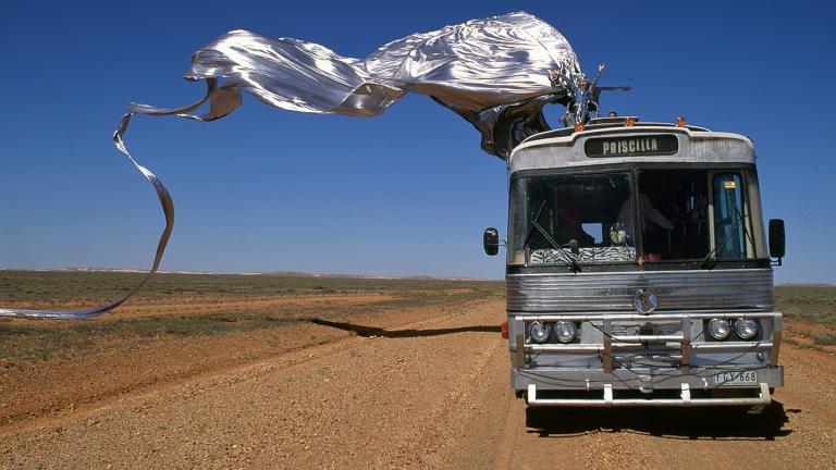 Guy Pearce in drag miming to an opera aria on top of a bus travelling through the desert