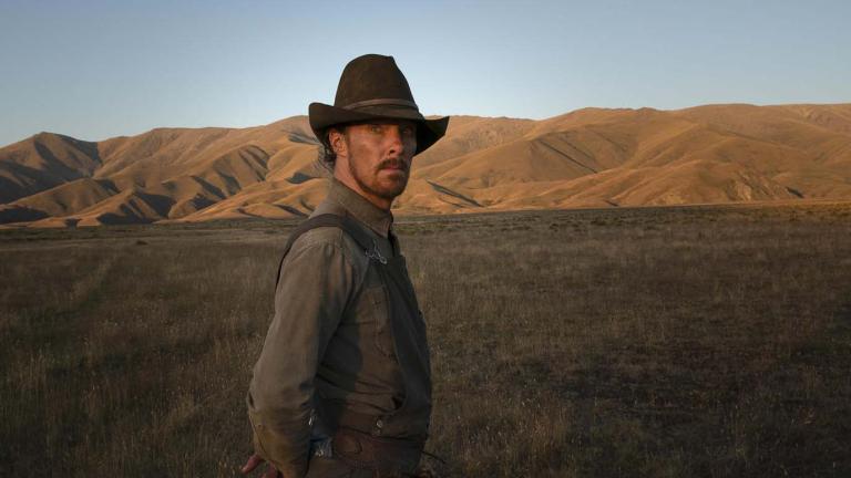 Benedict Cumberbatch dressed in weathered rancher's hat and overalls stands upright on his Montana farm, staring at the camera